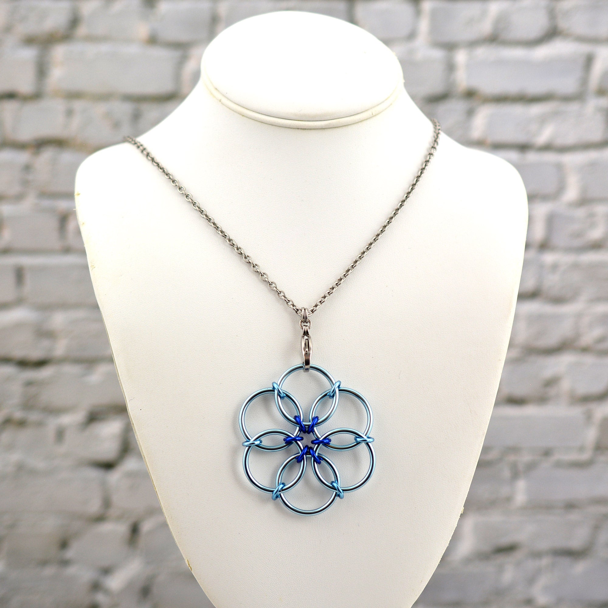 The Light Blue Fidget Flower necklace displayed on a white jewelry neck form against a white brick background. The pendant features light blue rings with a deep royal blue accent center.