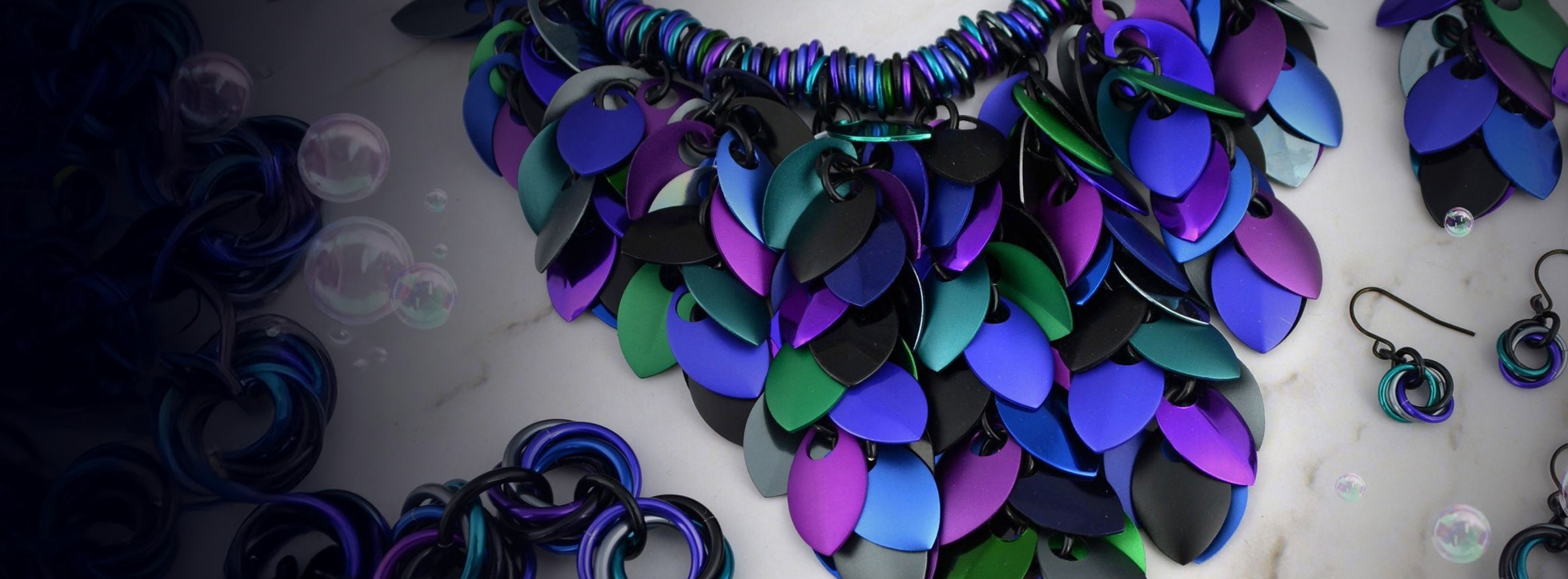 Statement scalemaille necklace in black, blue, purple and green against a white marble background.