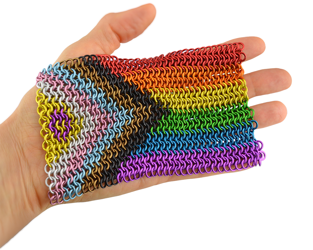 Artisan Rebeca Mojica's hand holding a vibrant, rectangular piece of chainmaille forming the intersex inclusive progress pride flag (gold triangle with a purple circle, chevron stripes in white, pink, light blue, brown and black, and the rainbow stripes).