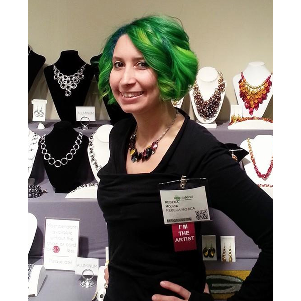 Rebeca Mojica, the chainmaille artist, smiling while standing in her jewelry display booth at the One of a Kind Show. She is wearing a black top and a colorful chainmaille necklace, with several other bold, intricately woven statement necklaces and earrings displayed on busts behind her, showcasing the variety of her award-winning chainmaille designs.