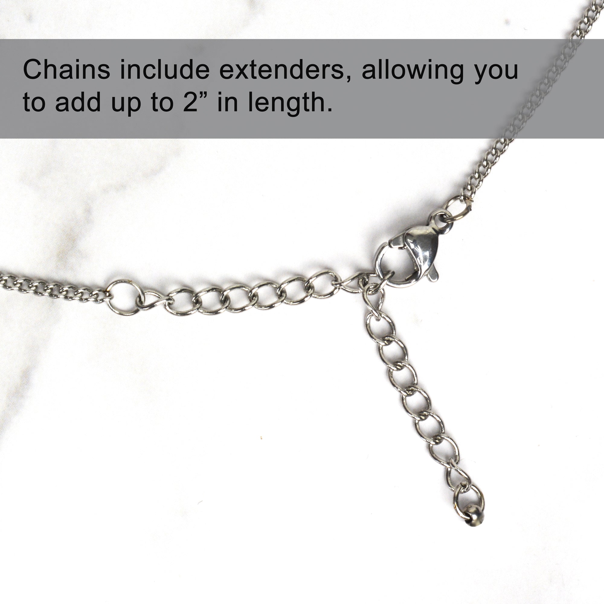 A close-up of a silver-toned necklace chain showing a lobster clasp and a 2-inch extender chain with visible oval links, on a white marble background. Text overlay states: "Chains include extenders, allowing you to add up to 2" in length
