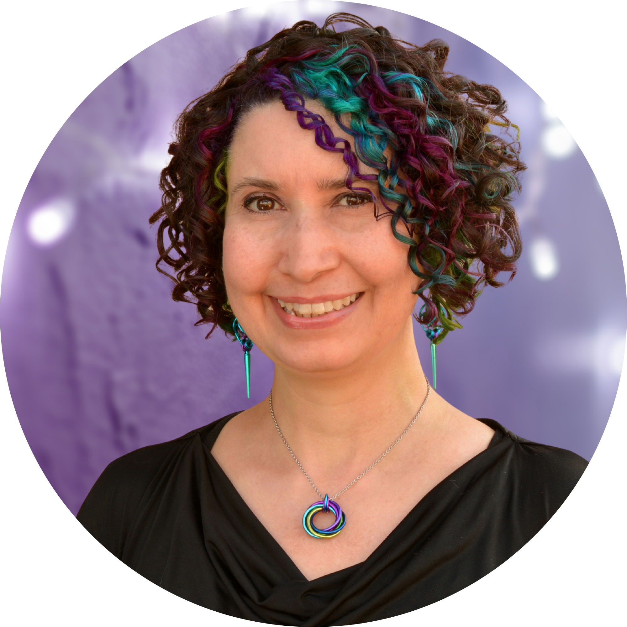 Headshot of Chainmaille artisan Rebeca Mojica, a light-skinned woman with a friendly smile, photographed in a circle against a soft purple background. Her naturally curly, short dark hair is accented with strands dyed in bright teal, magenta, and lime green. She wears a black top, a Peacock Swirl chainmaille pendant, and long, spiked dice chainmaille earrings, embodying the brand's focus on color and authentic self-expression.