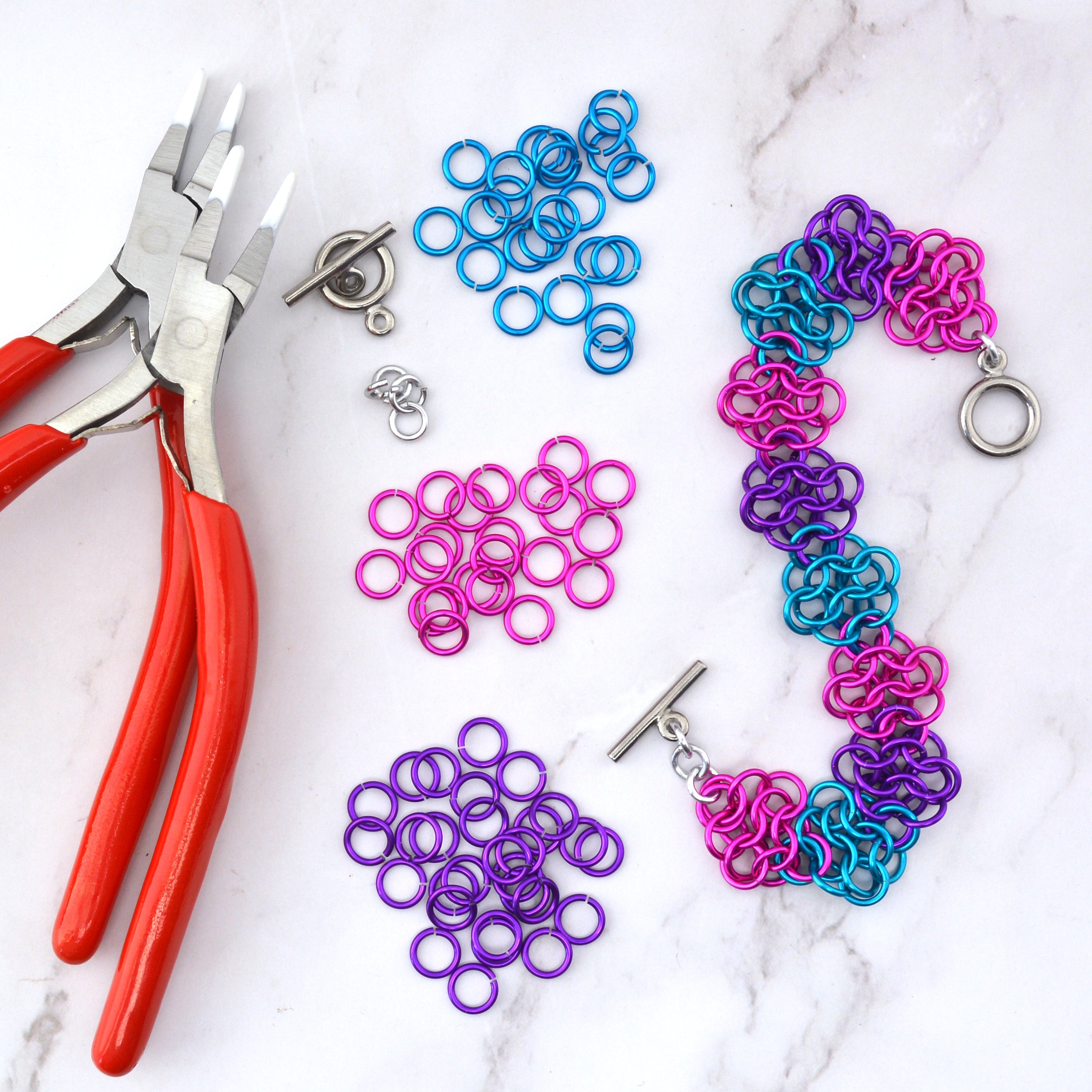 Colorful chainmaille bracelet in the Rosettes weave using hot pink, violet and turquoise jump rings. The bracelet is arranged in an S shape, alongside pliers and piles of loose jump rings on a marble surface
