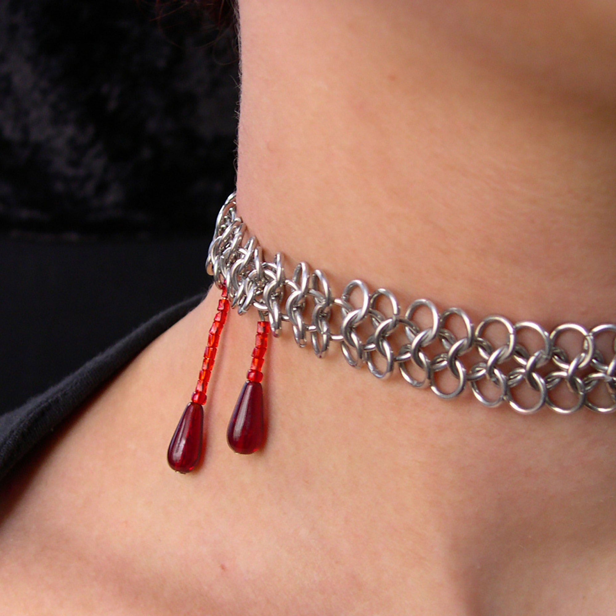 Closeup of neck with silver color chainmaille mesh band and two deep red drops meant to look like blood trickles. The red drops are positioned on the side of the neck, to allude to a vampire bite.