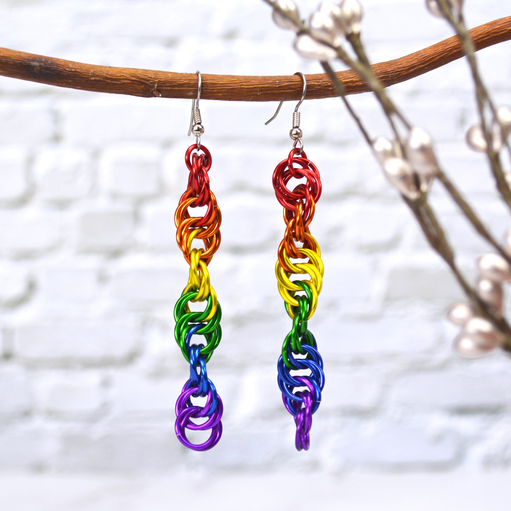 Rainbow chainmaille earrings in a spiral weave. Earrings hang on a branch with a blurred background