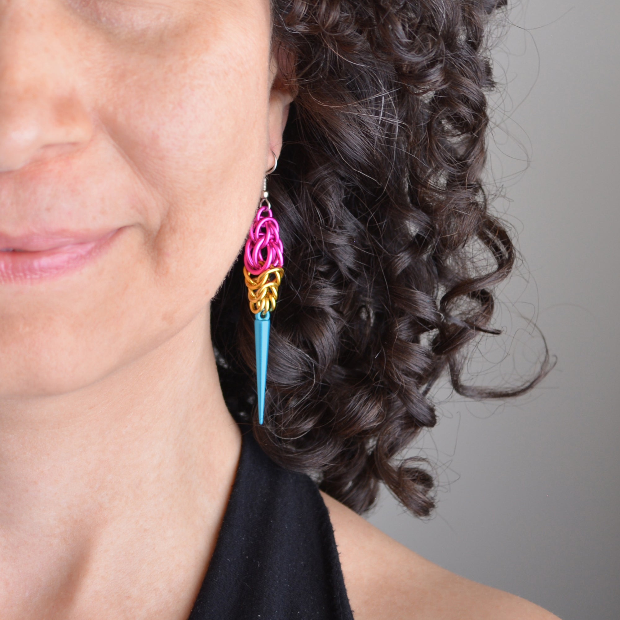 Closeup of the Pansexual Pride Spike earrings being worn. The chainmaille earrings are pink, gold and turquoise to best match the pansexual pride flag colors. Only the bottom quarter of the person's face is shown; they have curly brown hair behind the earring.