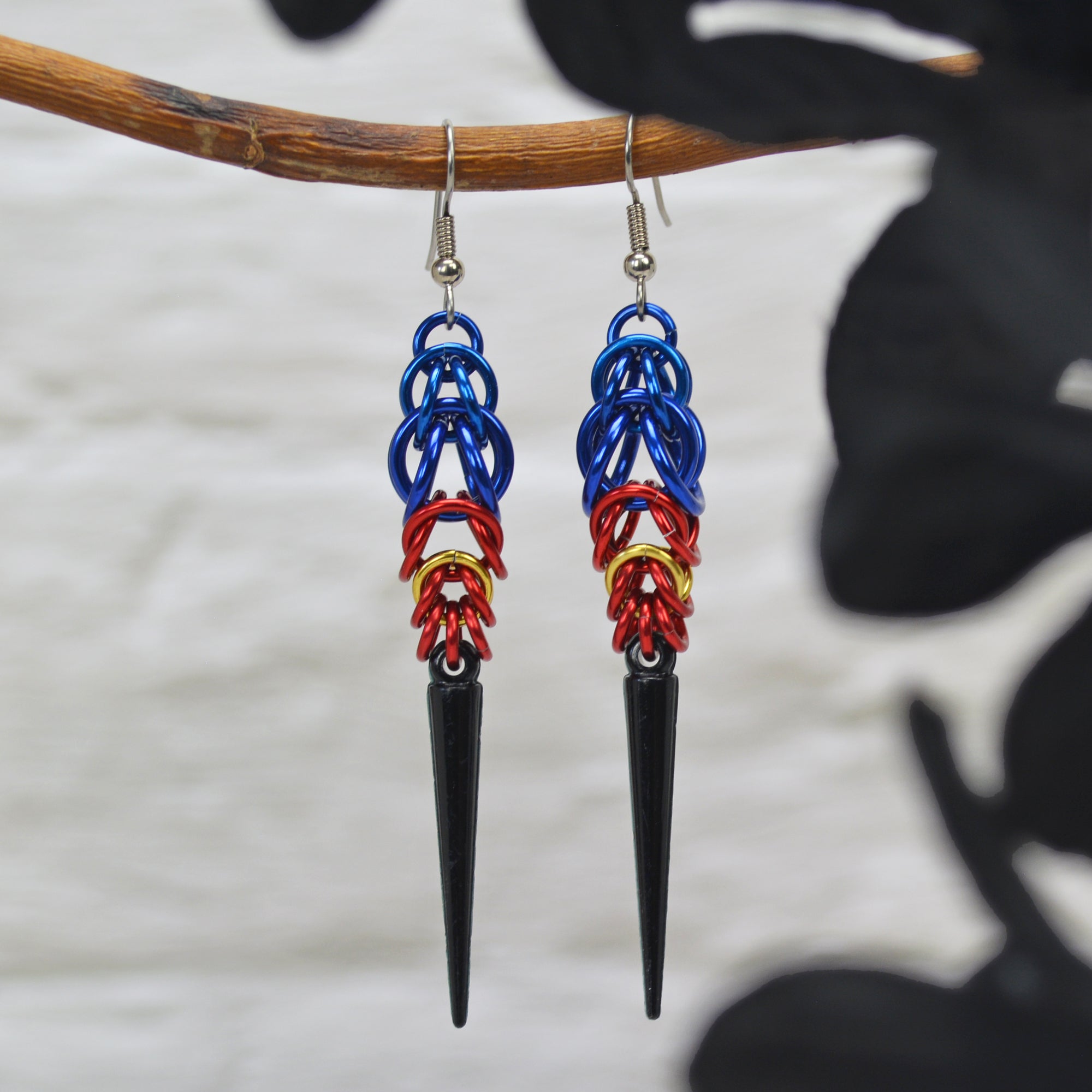 Poly pride chainmaille earrings hanging from a grey branch with a blurred white brick background. Earrings are blue at the top in the Full Persian weave, followed by red and gold links in the Box weave, with a black acrylic spike at the base.