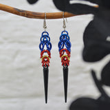  Poly pride chainmaille earrings hanging from a grey branch with a blurred white brick background. Earrings are blue at the top in the Full Persian weave, followed by red and gold links in the Box weave, with a black acrylic spike at the base.