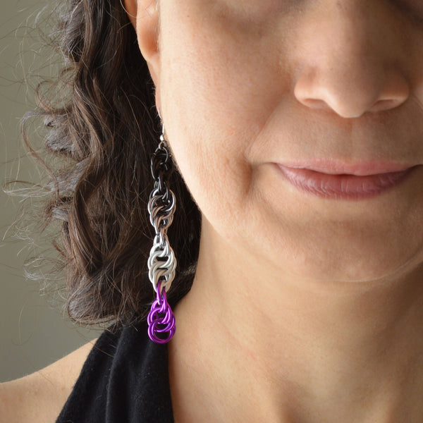Closeup of the bottom right quadrant of a woman’s face wearing a long chainmaille spiral earring in the ace pride colors - black, grey, white and violet