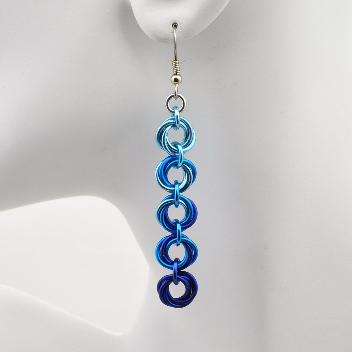 Another closeup of chainmaille earrings on display head. Earrings are 5 knots linked vertically and transitioning from a top knot of light blue to a bottom knot of dark blue. Five Knot Earrings by Rebeca Mojica