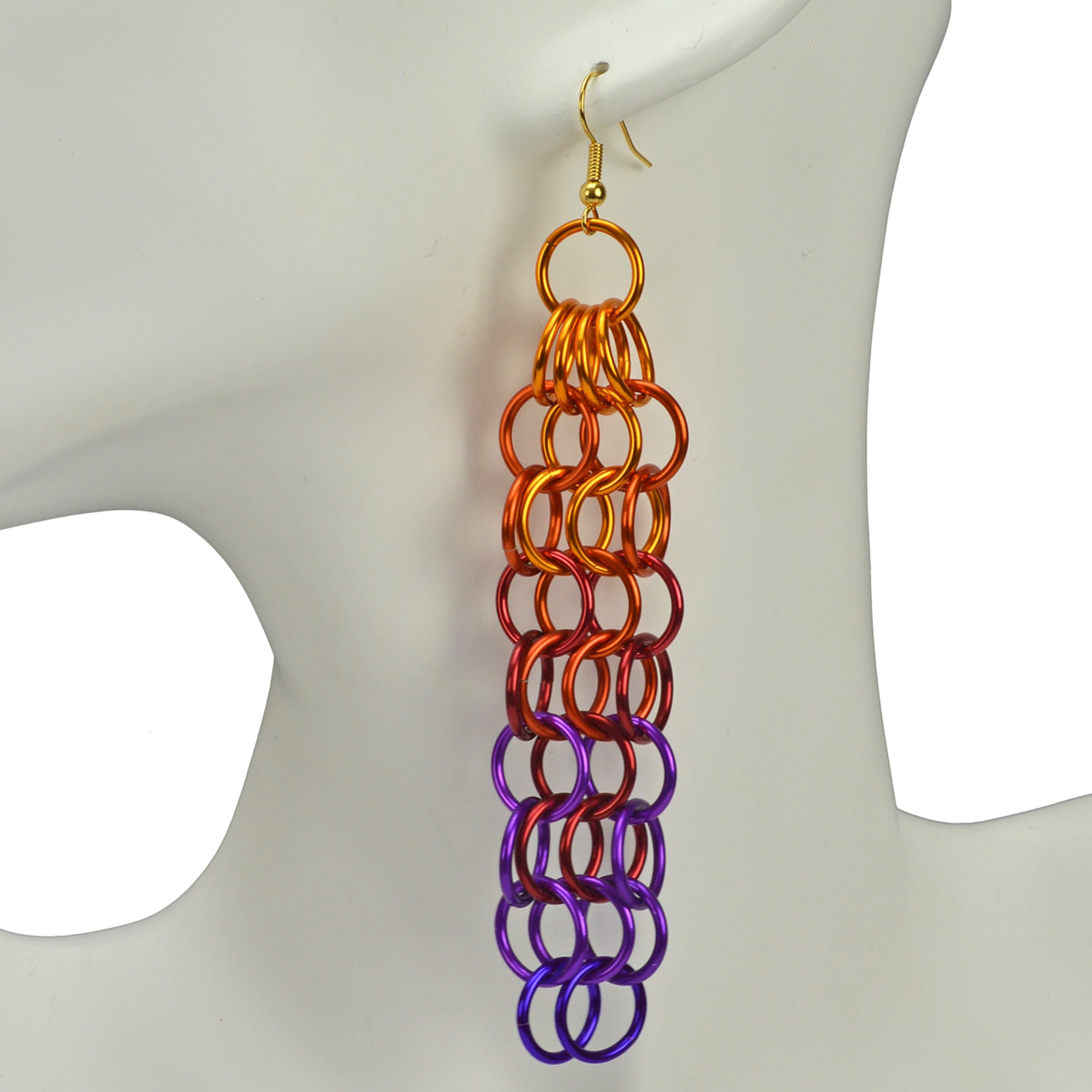 Closeup of Rebeca Mojica's long mesh chainmaille earrings in the sunset ombre. The earrings have a fiery look with orange at the top, transitioning to red, violet and purple. The earrings alternate rows of 4 links with rows of 3 links.