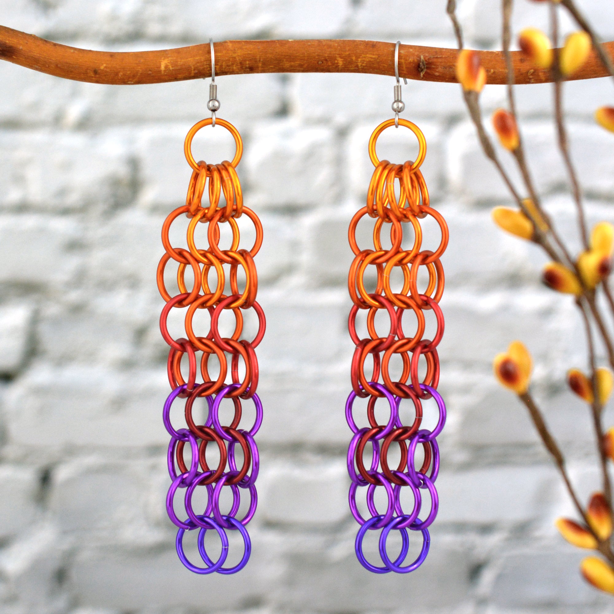Large link chain mesh earringsin a fiery ombre hanging from a branch. The long chainmaille earrings are light bright orange at the top, red in the middle, transitioning to violet and purple at the bottom. The background is a blurred light grey brick wall.