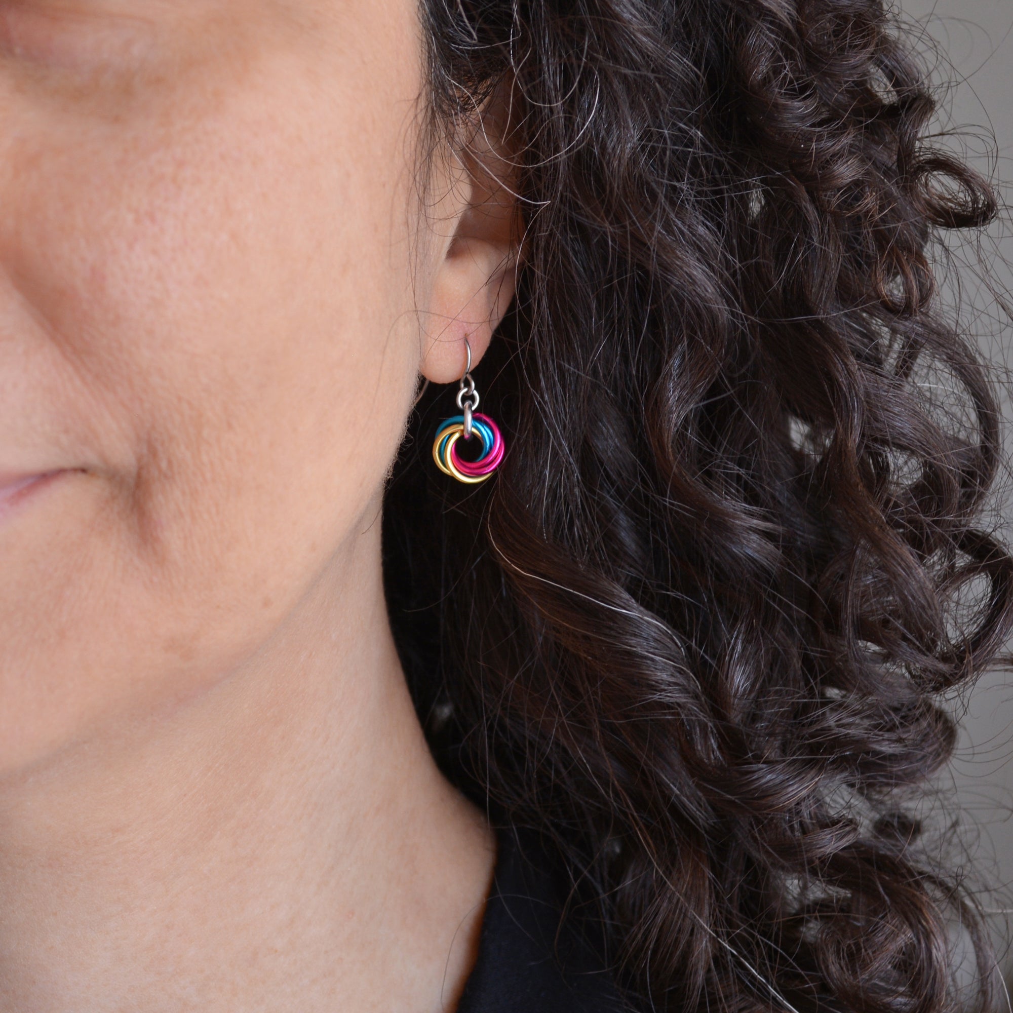 A close-up shot of chainmaille artisan Rebeca Mojica’s ear and the side of their face, showing a Pansexual flag mini knot earring (pink, yellow, blue links) being worn. Rebeca has dark, curly hair.