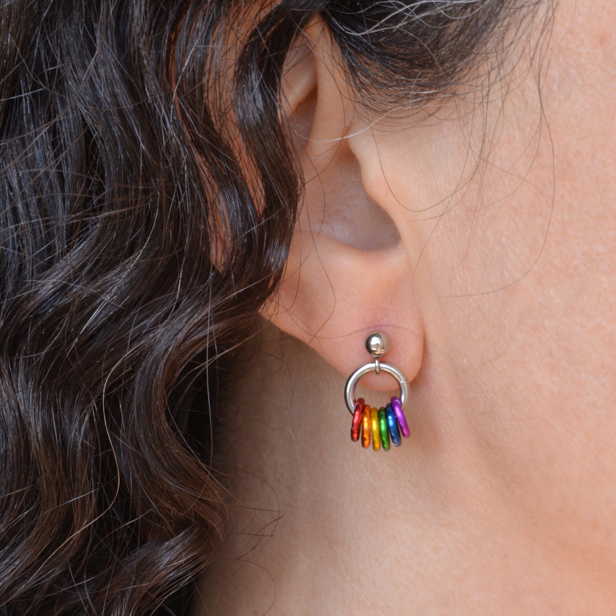 Closeup of a tiny chainmaille earring in LGBTQ rainbow flag colors. Earring is shown on the earlobe of a person with dark brown and grey curly hair. Earring has a small stainless steel ball at the top of the post, with a medium size steel ring hanging from the ball. Small anodized aluminum jump rings hang from the medium steel ring - from left to right the colors are red, orange, yellow, green, blue and violet.