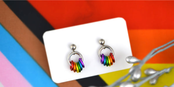 Tiny chainmaille rainbow pride stud earrings on a small white card set on a background of the top portion of a progress pride flag. Earrings by artisan Rebeca Mojica image