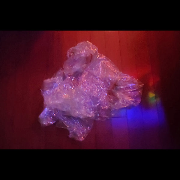 A close-up photograph of a crumpled, translucent plastic bag sitting on a dark wood floor, illuminated by a warm red light from above and a blue-purple light from the side.