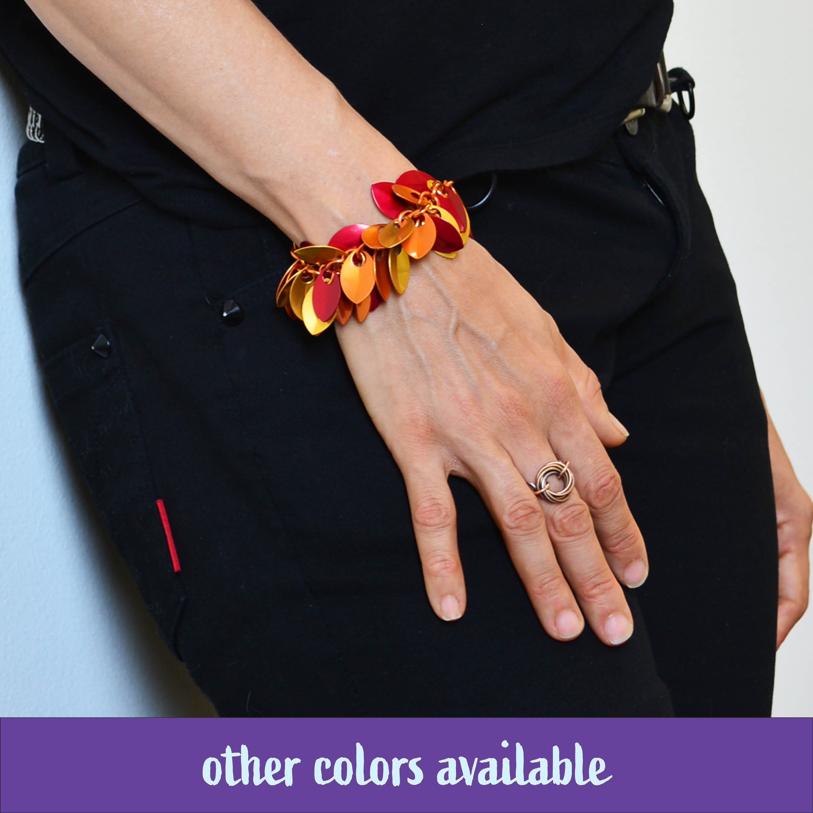 Close-up of artisan Rebeca Mojica's arm wearing the Cascading Leaves Bracelet in the Flame colorway, featuring shades of red, orange, and gold scales. She is wearing black pants and a black top. A banner reads, "other colors available."