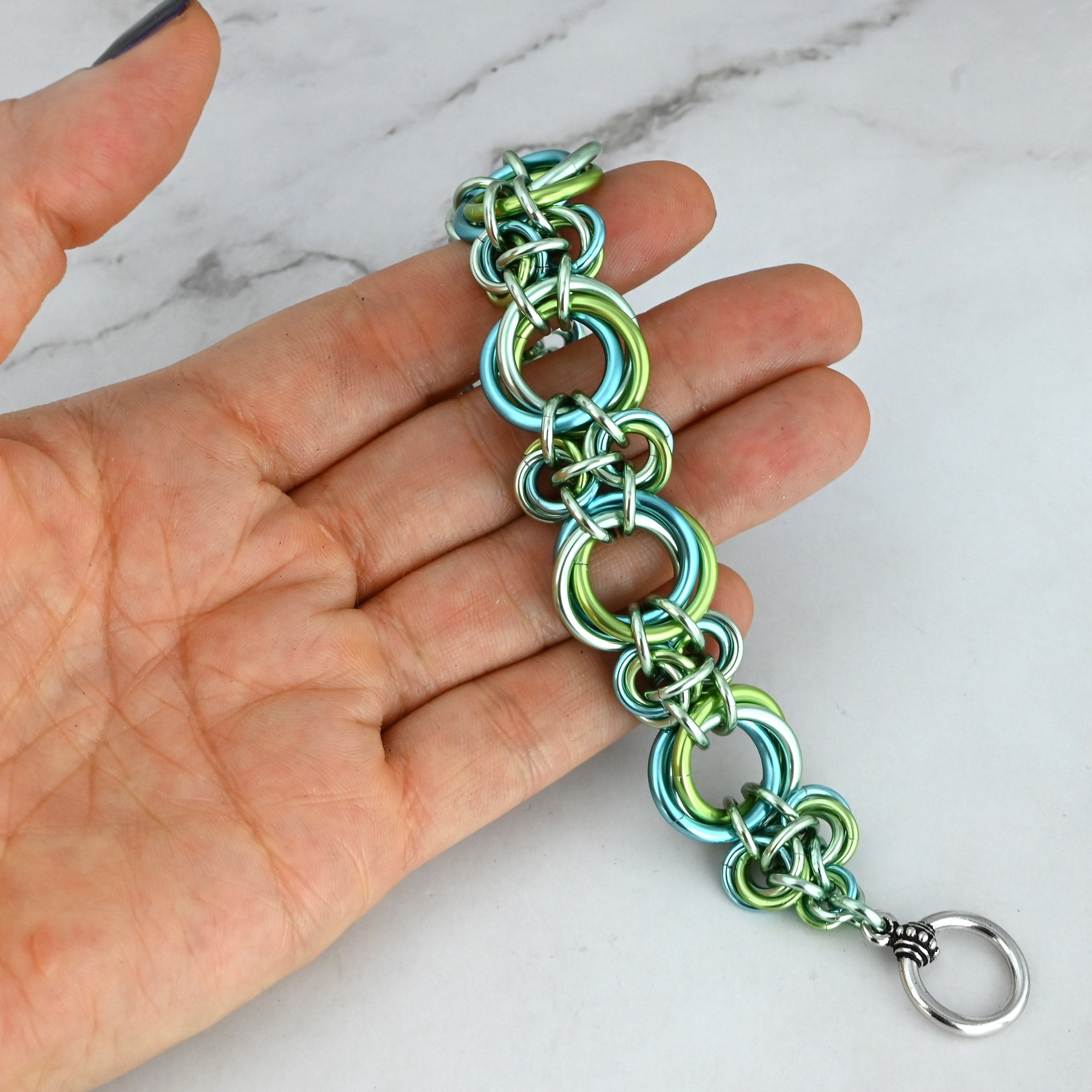 A close-up view of the "Absinthe" chainmaille bracelet draped across a person's fingertips. The bracelet rests diagonally across the fingers, clearly illustrating the staggered, 3D texture and the alternating structure of the doubled small knots and the tri-color large rings.