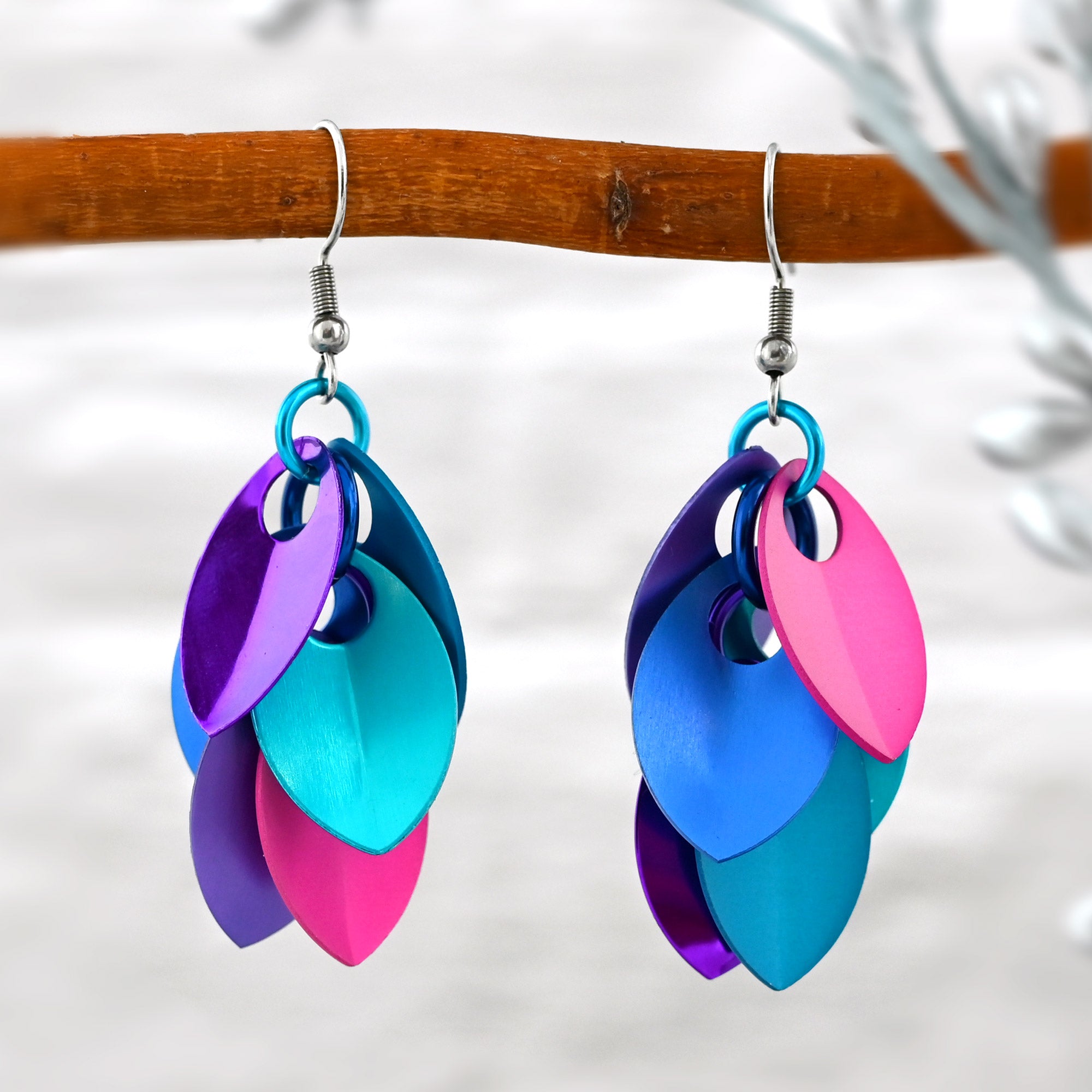 Close-up of a pair of hot pink, blue, and violet aluminum cascading leaves earrings hanging from a branch against a blurred white brick background.