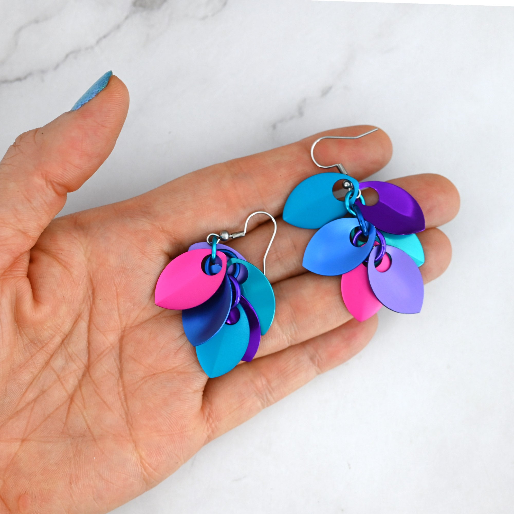 A pair of small scalemaille earrings resting in a person’s fingertips to demonstrate their mismatched design in a vibrant blend of blues, purples and hot pink.