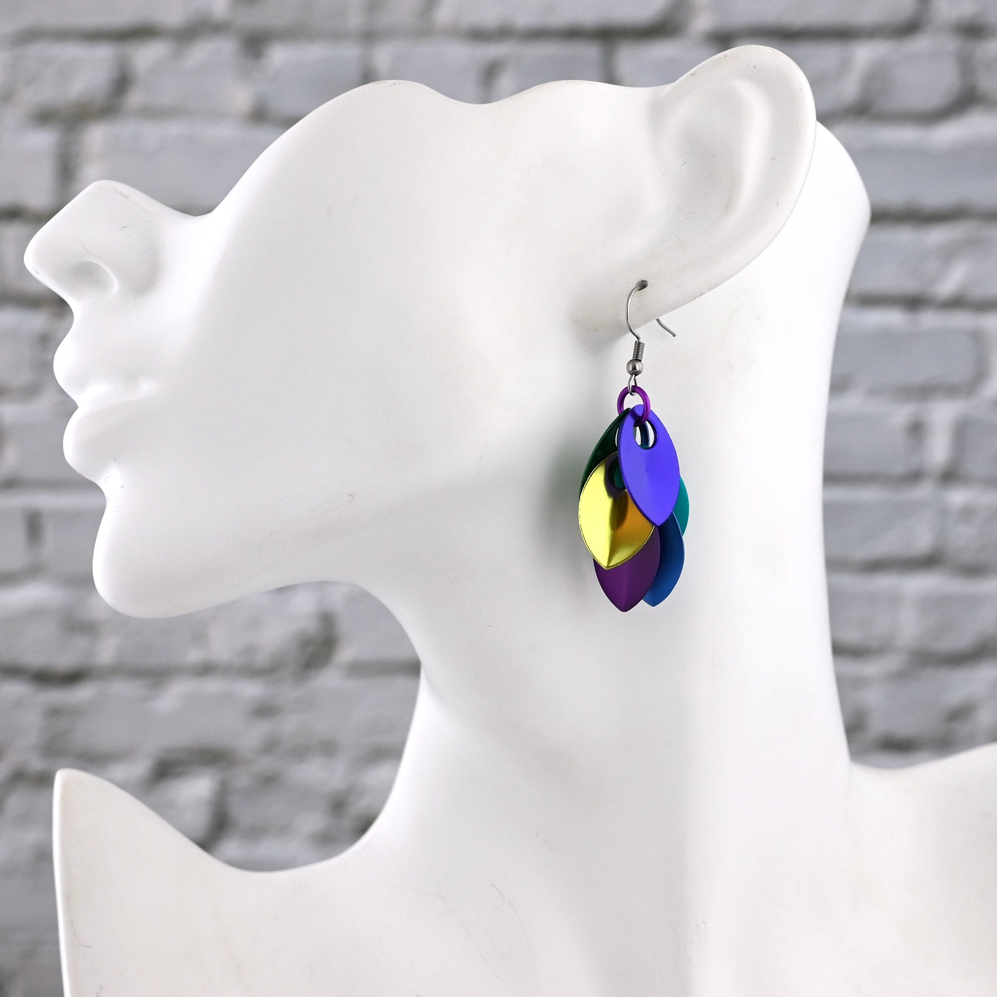 Multicolored scalemaille earrings on a white mannequin ear against a gray stone wall background