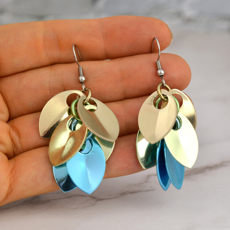 Chainmaille artist Rebeca Mojica holding a pair of seashell ombre dangle earrings, showing the hand-linked aluminum leaves (scales).