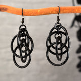 Black intertwined hoop chainmaille earrings hanging from a wooden branch, highlighting the glossy finish of the rings.