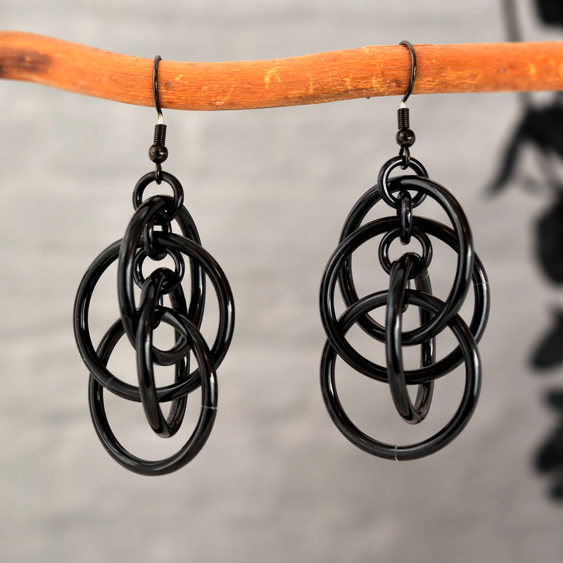 Black intertwined hoop chainmaille earrings hanging from a wooden branch, highlighting the glossy finish of the rings.