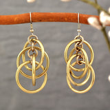 A close-up of champagne-colored intertwined hoop chainmaille earrings hanging from a wooden branch, highlighting the neutral color finish and sheen.