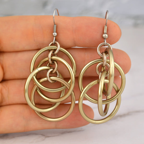A close-up shot of the champagne-colored intertwined hoop chainmaille earrings being held by Rebeca Mojica's fingertips, providing a sense of their scale and lightweight feel against a marbled background.