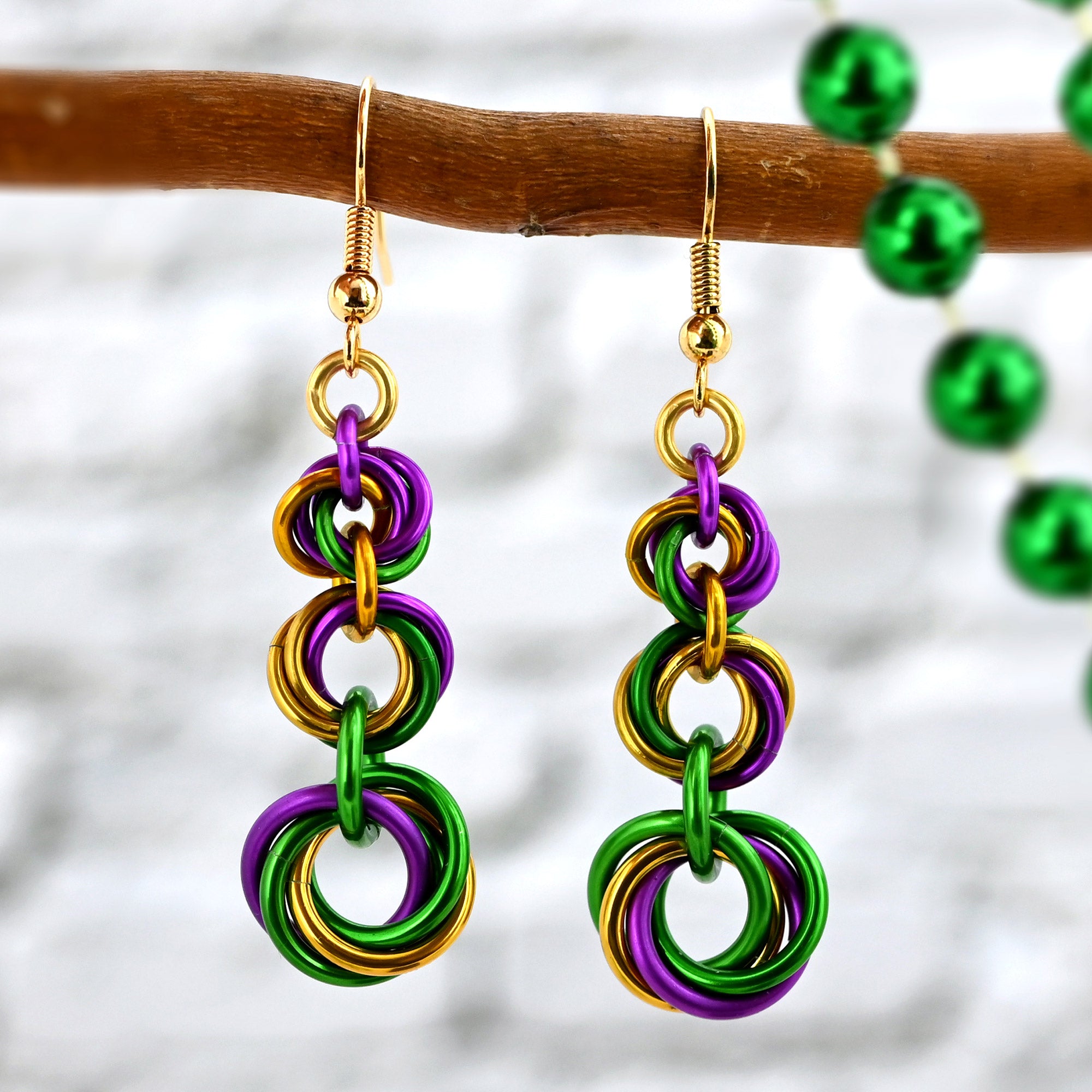 Close-up of two graduated chainmaille knot earrings hanging from a natural wooden branch. Each earring has 3 knots that increase in size from top to bottom, and each swirl has a mixture of violet, green and gold links.