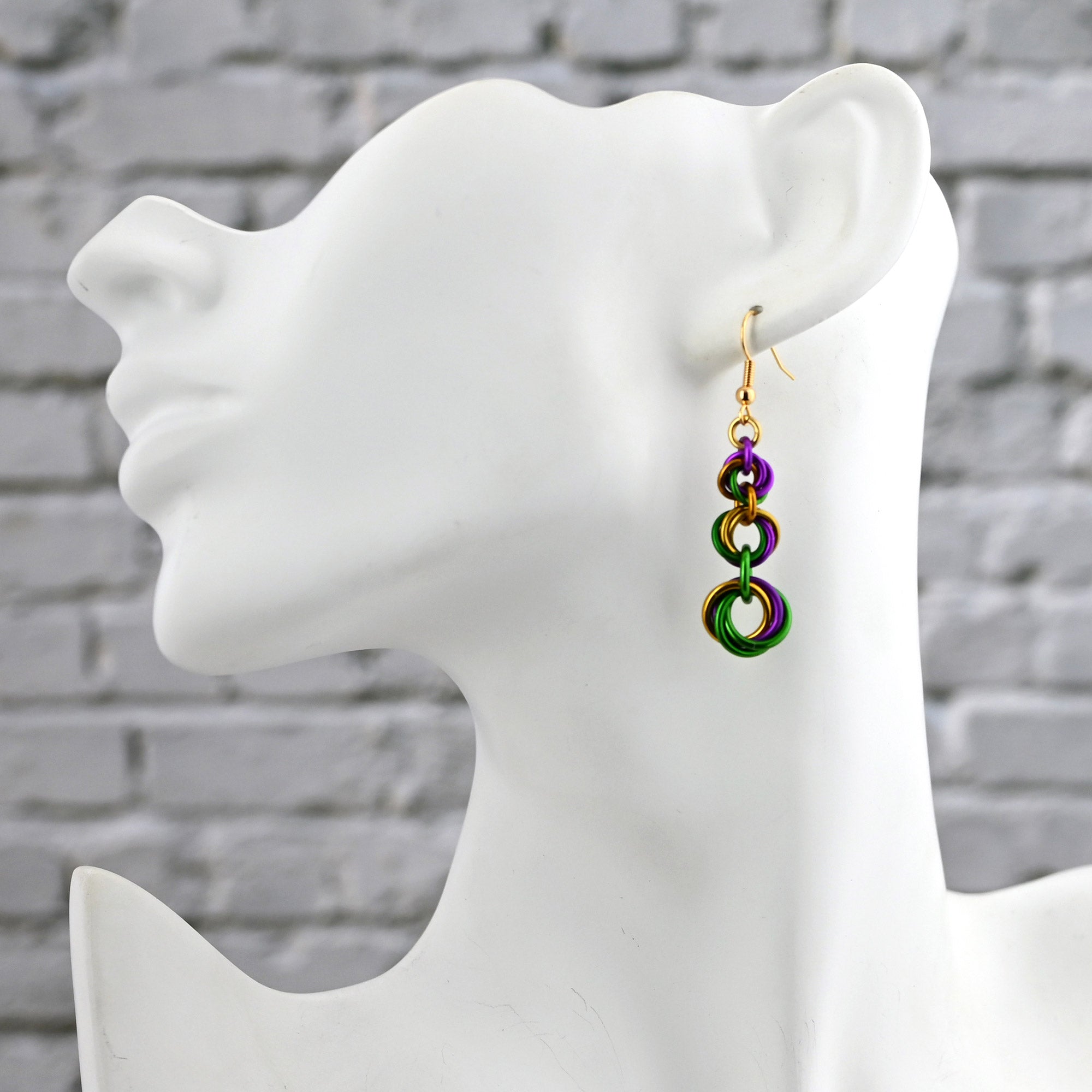 Side view of a white mannequin bust wearing a graduated chainmaille knot earring to show how the purple, green, and gold colors sit against the neck.
