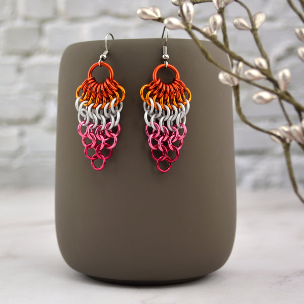 A pair of chainmaille mesh earrings hanging from the top of a grey mug. The earrings are dagger shaped wide at the top tapering down to a single link at the bottom row, in the colors of the lesbian pride flag—dark orange, bright orange, white, pink and fuchsia. Background is a off-white brick wall and marble floor.