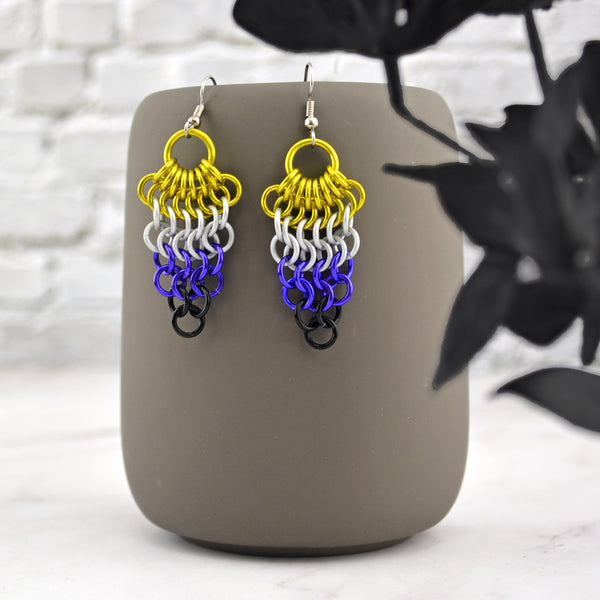 A pair of chainmaille mesh earrings hanging from the top of a grey mug. The earrings are dagger shaped wide at the top tapering down to a single link at the bottom row, in the colors of the nonbinary pride flag—yellow, white, purple and black. Background is an off-white brick wall and marble floor.