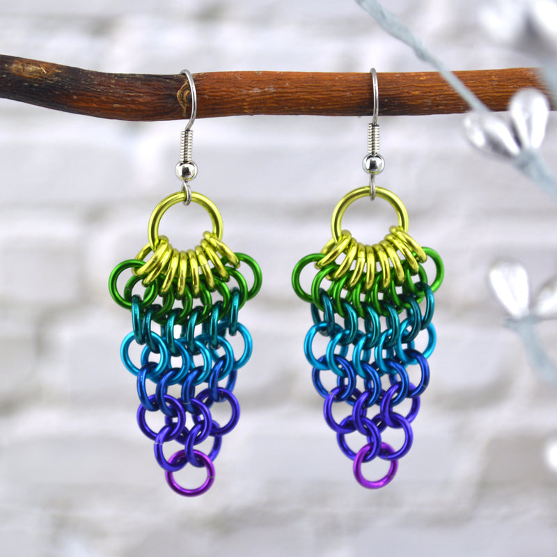 A pair of chainmaille mesh earrings hang from the top of a branch. Earrings are reverse teardrop shape with a large chartreuse ring at top. From that ring, a row of 8 smaller chartruese rings hang and are connected to a row of 7 green rings beneath them. Each row has fewer rings: 6 teal rings, 5 turquoise rings, 4 blue rings, 3 purple rings, 2 purple rings and 1 violet ring.
