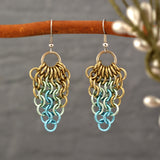 A pair of metal mesh teardrop shaped earrings are shown hanging from a thin, curved wooden branch against a blurred gray background. The photo highlights the delicate, chainmaille weave and pastel colors in champagne, seafoam and light blue.