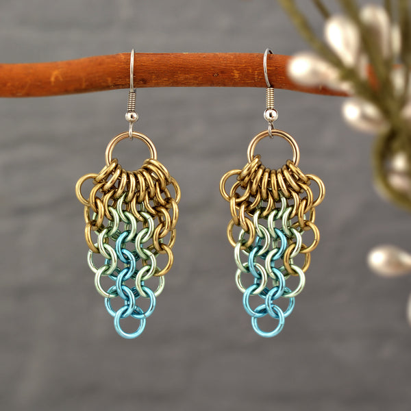 A pair of metal mesh teardrop shaped earrings are shown hanging from a thin, curved wooden branch against a blurred gray background. The photo highlights the delicate, chainmaille weave and pastel colors in champagne, seafoam and light blue.