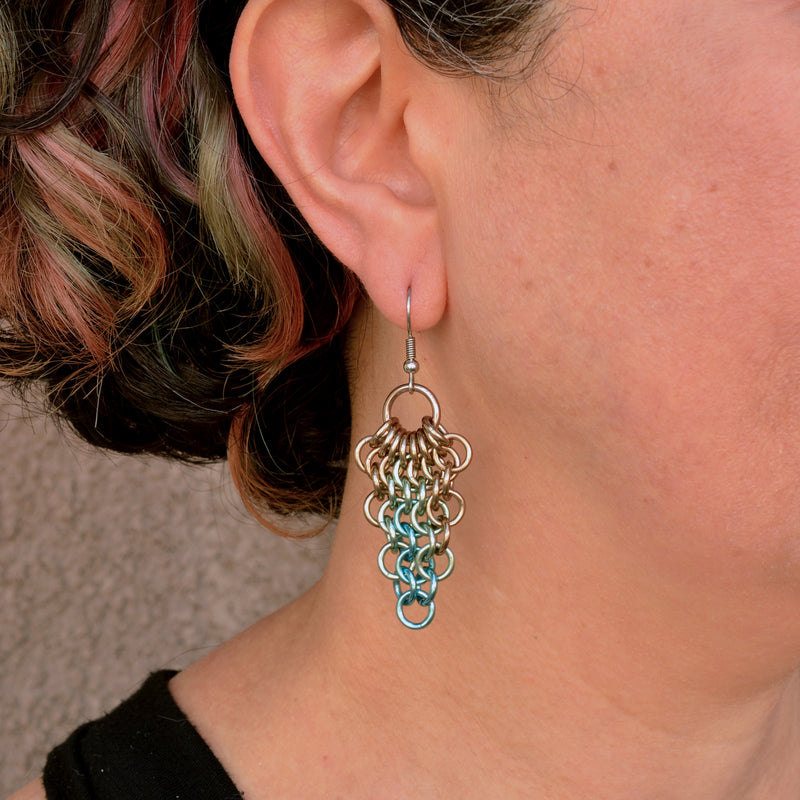 Artisan Rebeca Mojica wears a single seashell color teardrop mesh earring. The earring hangs just below her earlobe, showing its length and how it looks when worn. She has short, dark hair.