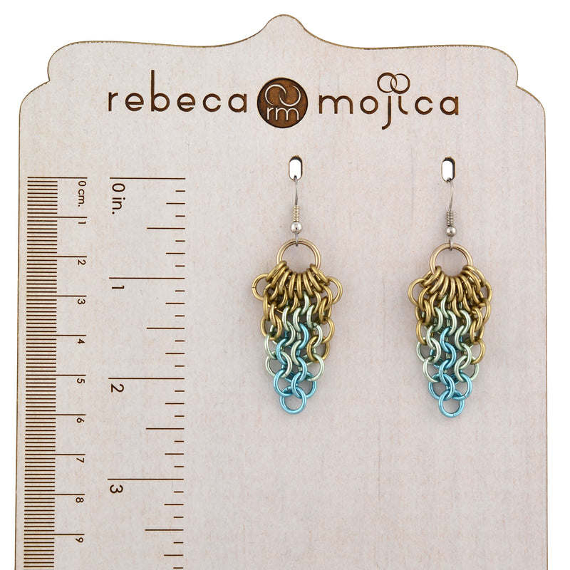 A pair of chainmaille mesh earrings are displayed on a wooden jewelry display with a ruler printed on it, showing the earrings are approximately 2.25 inches long. The brand name, "rebeca mojica," is visible at the top of the card.