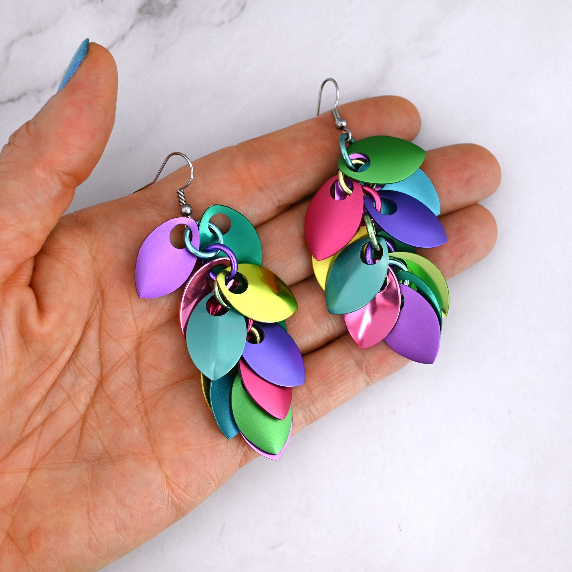 Saturated pastel earrings in a person’s fingertips to show the size and colors. The earrings are scalemaille, featuring pairs of mismatch anodized aluminum leaves that splay outward from a central chain of colorful jump rings. The leaf-shape scale components are green, pink, purple, blue and yellow.  