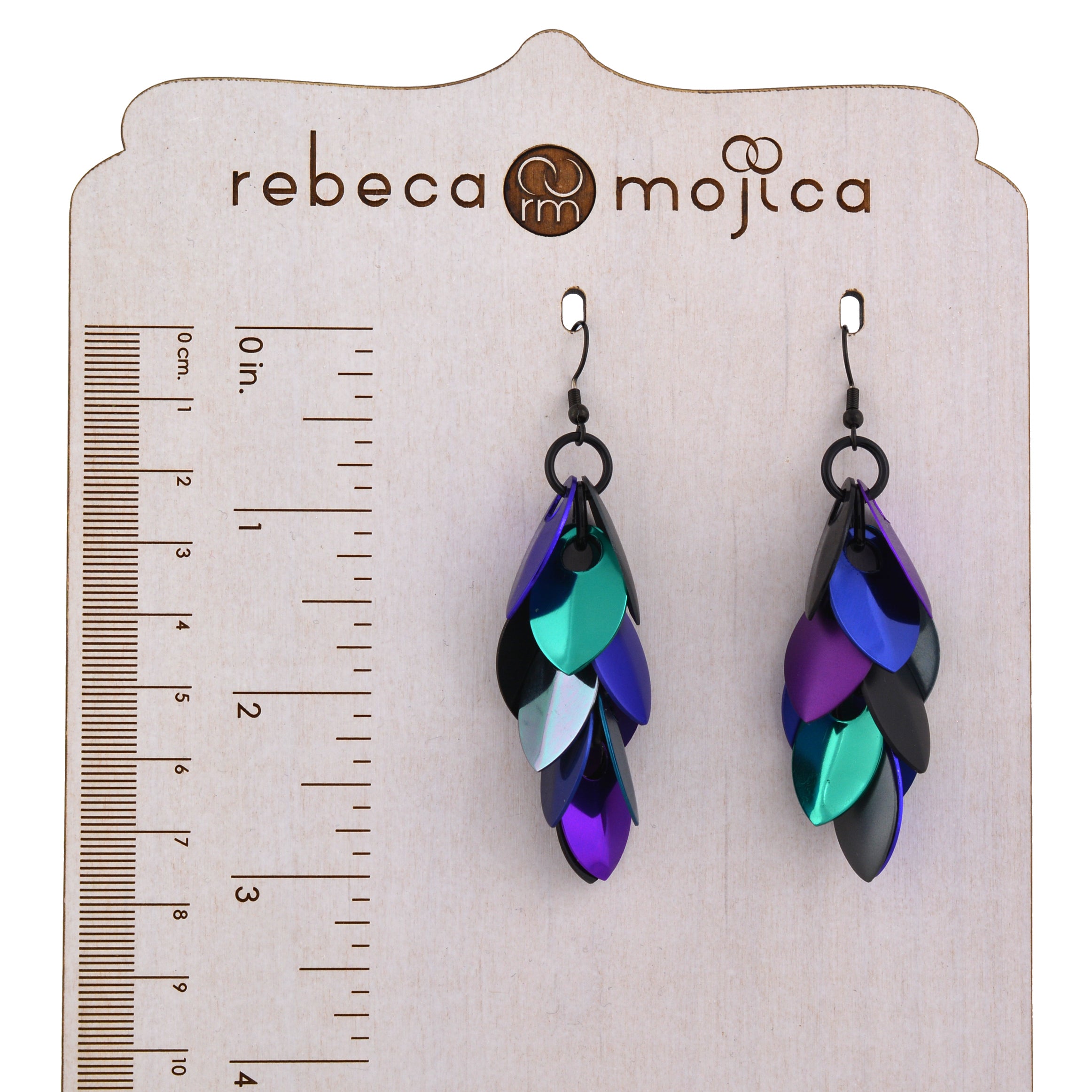 A pair of Rebeca Mojica's long, multicolored "Dark Mermaid" chainmail scale earrings hanging on a wooden display board next to a ruler, clearly showing the length is approximately 3 inches.