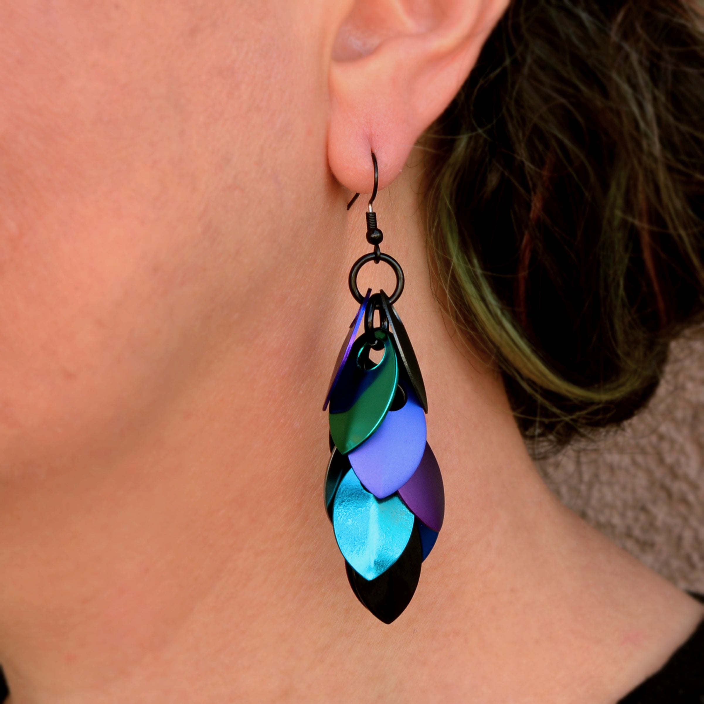 Close-up of artisan Rebeca Mojica's ear, highlighting the intricate detail of one "Dark Mermaid" scale earring with its shifting colors and black ear wire.
