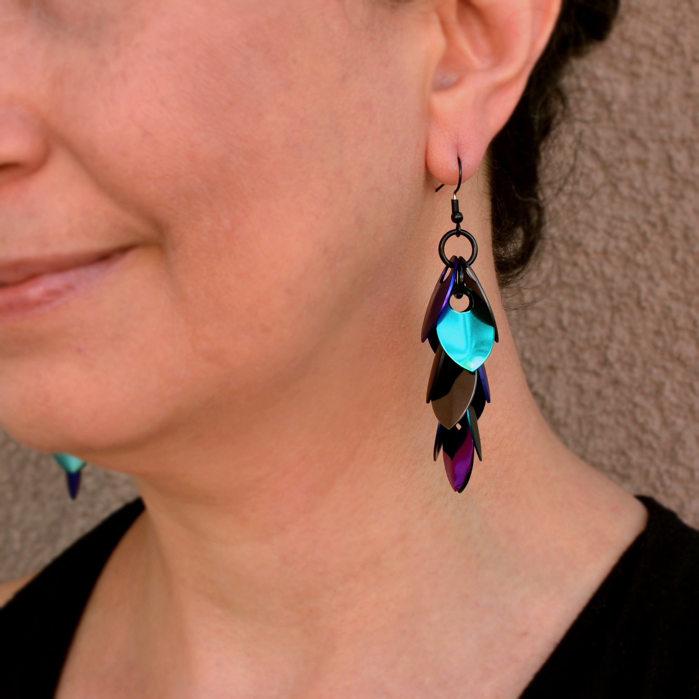 Artisan Rebeca Mojica models one long, iridescent Dark Mermaid feathered scalemaille earring, showcasing the mix of blue, teal, purple, and black scales as it drapes beside her neck.