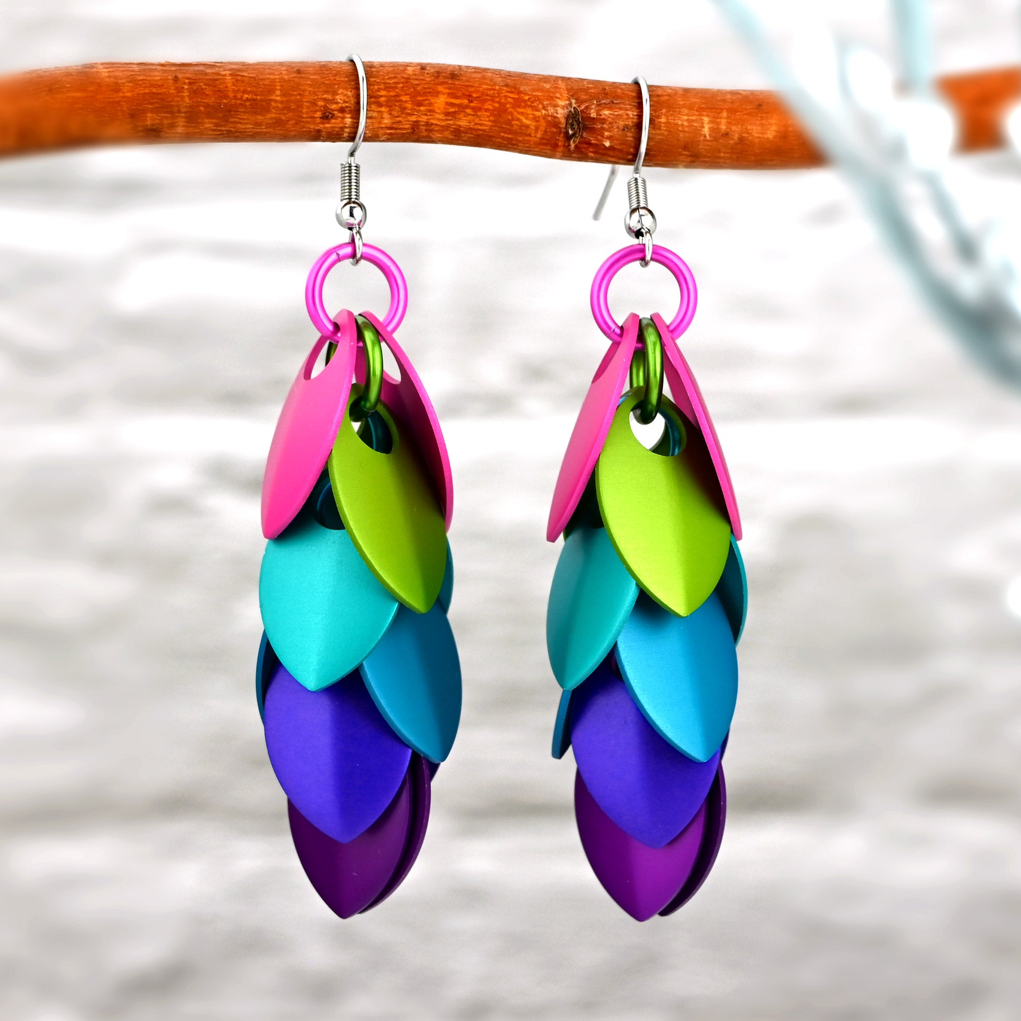 Close-up of vibrant long feathered earrings hanging from a branch against a blurred white brick background. From top to bottom, the earrings have overlapping pairs of hot pink, chartruese, aqua, azure, purple and violet scales linked with a single strand of jump rings in the same colors.