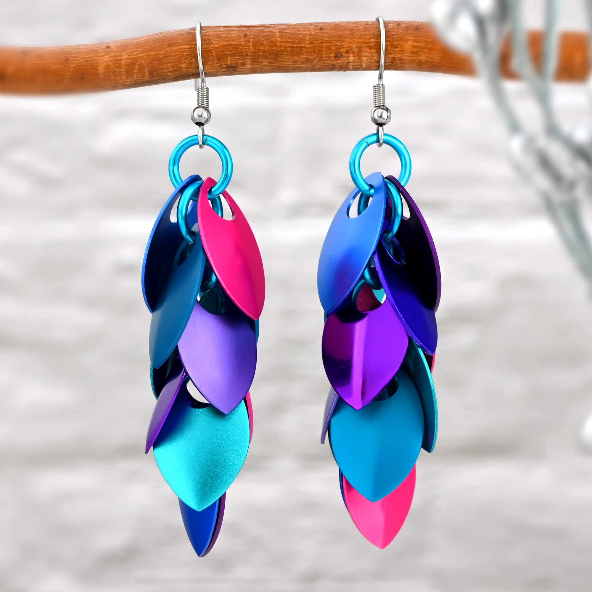 Close-up of a pair of long aluminum scalemaille earrings in pink, blue, and violet hanging from a branch, highlighting the mix of matte and shiny finishes and the vibrant colors.