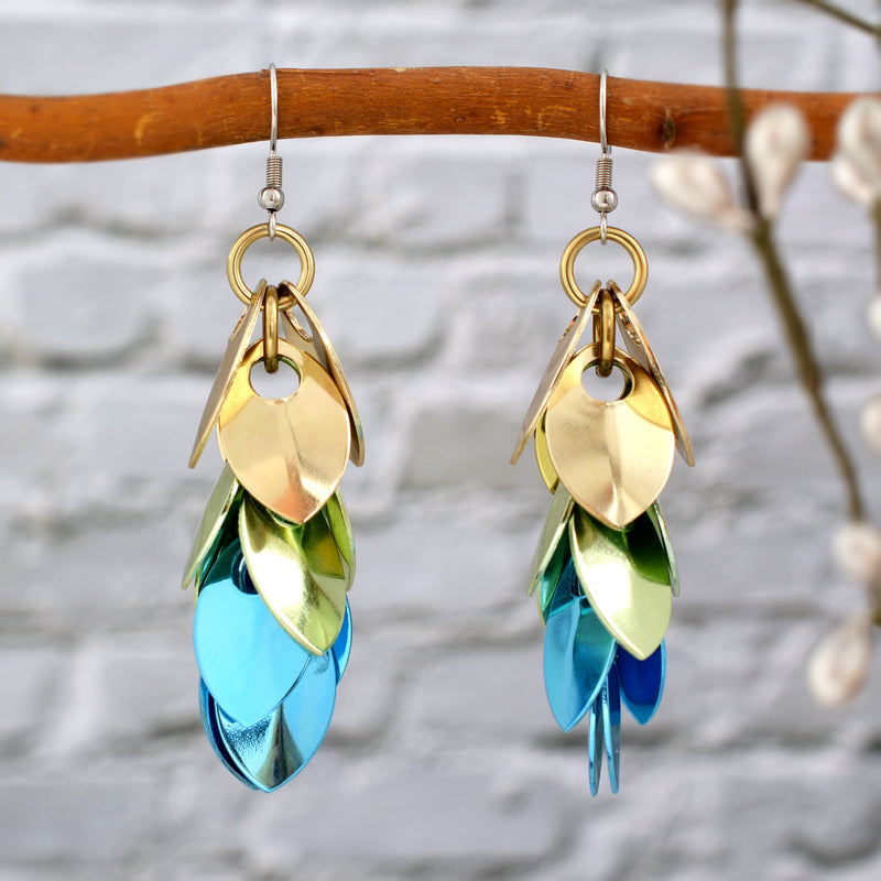 Rebeca Mojica's Cascading Leaves scalemaille earrings hang from a brown branch against a light brick wall. The shiny champagne, seafoam, and light blue scales reflect light, making the earrings stand out.
