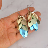 Artisan Rebeca Mojica holds the pair of earrings, showcasing the full length and the way the metallic colors shimmer as they hang. A white marble surface is in the background.