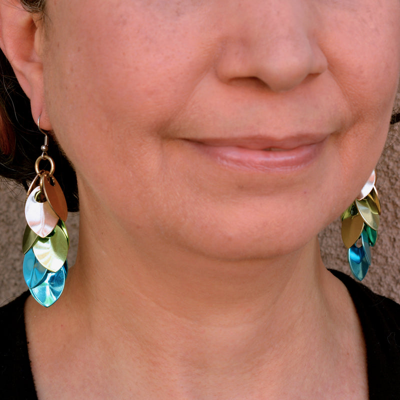 A photo of artisan Rebeca Mojica wearing the long, elegant scalemaille earrings in champagne, seafoam and blue colors, showcasing how the “leaves” (scales” reflect the light
