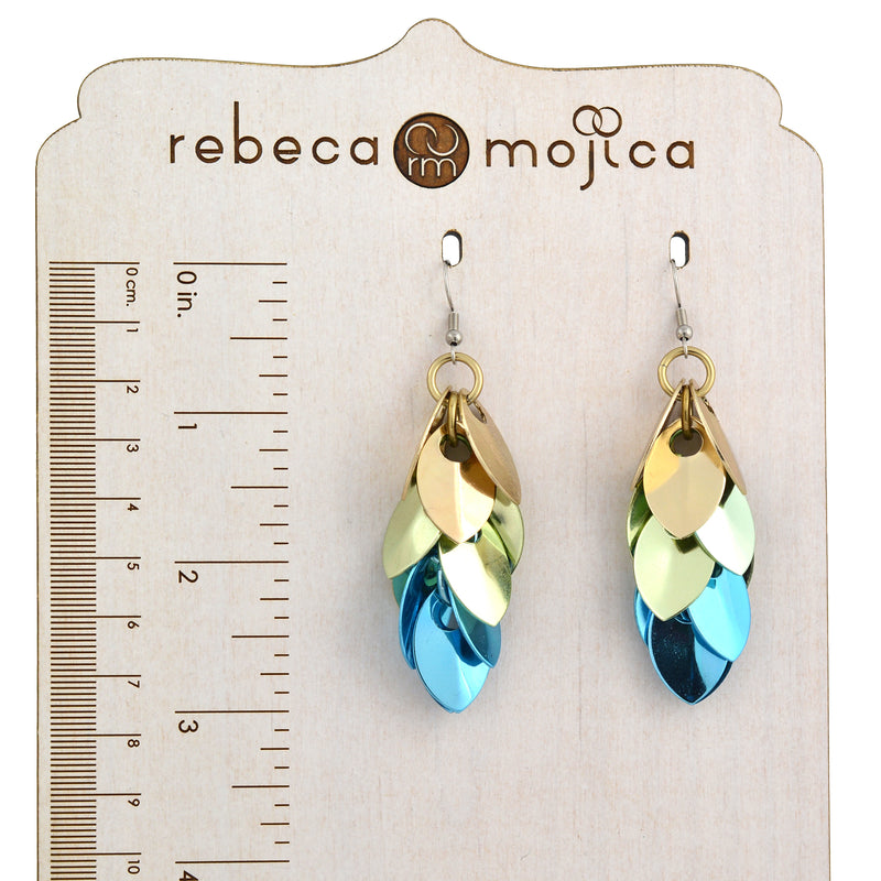 A pair of elegant scalemaille earrings in champagne, seafoam, and light blue are displayed on a light-colored wooden earring display stand with the brand name "rebeca mojica" and a size ruler on the left side showing the earrings measure about 3” long (7.6cm)