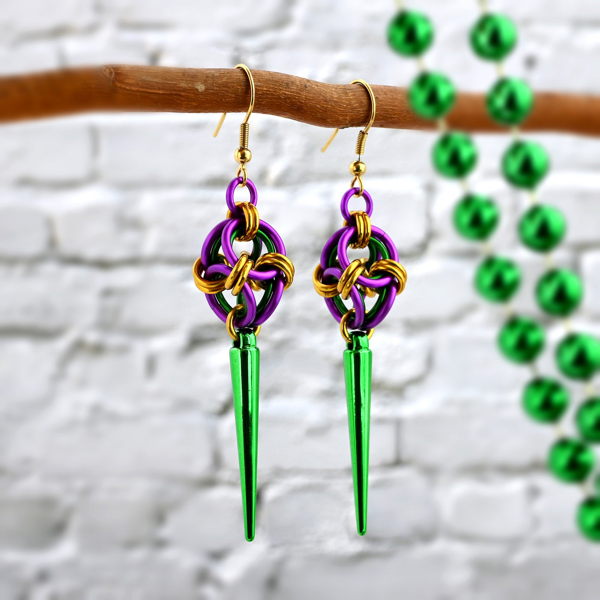 Angled view of Mardi Gras polyhedron spike earrings hanging on a branch with blurred green carnival beads in the background.